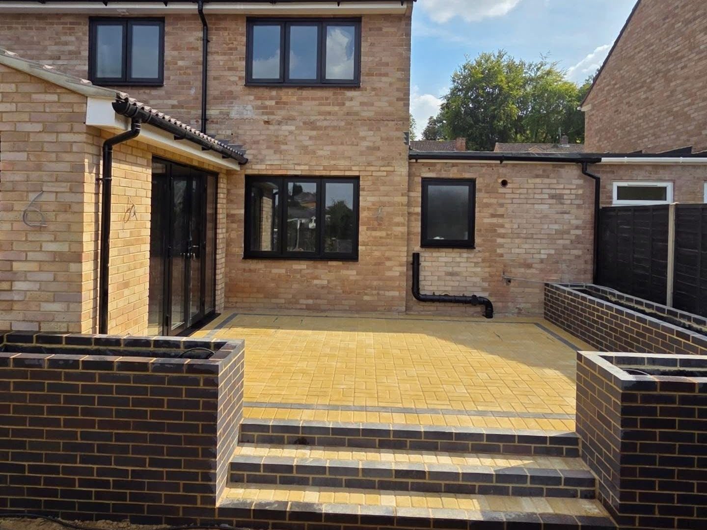 The same garden with a raised buff block paved patio, brick walls and charcoal steps